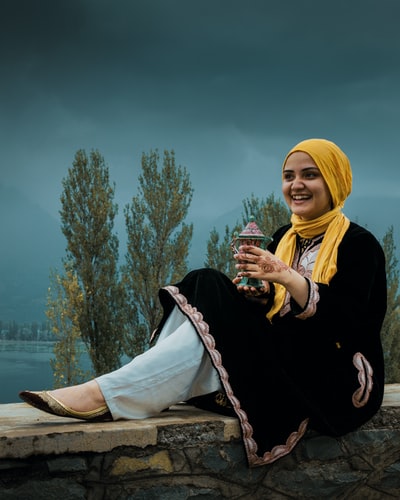 During the day, a man in a black jacket, wearing a white turban women sitting on a brown wood
